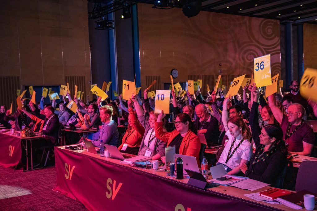 Bilde av delegater på landsmøtet som holder delegatskilt i lufta.